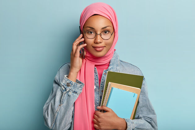 阿拉伯语人 科技 种族 沟通观念戴着传统头巾的漂亮女孩与团友通电话 讨论未来项目 拿着两个螺旋笔记本 在室内摆姿势围巾高兴伊斯兰