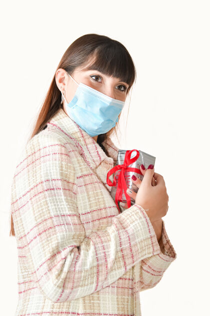 家庭一个年轻女子的特写镜头 她戴着一个外科口罩 把一个礼盒放在胸前 背景很浅面具年轻女性圣诞礼物