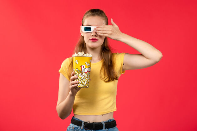 肖像前视图年轻的女性在电影院拿着爆米花在-d太阳镜在浅红色的墙上电影院小吃有趣的电影剧院女性爆米花