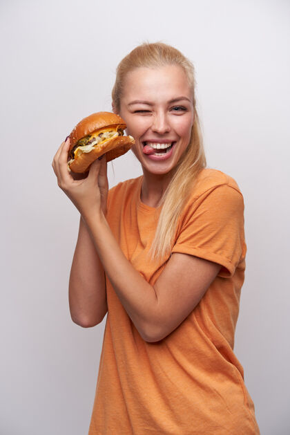 垃圾有趣的镜头 可爱的金发女郎扎着马尾辫 对着镜头眨眼 露出舌头 手里拿着新鲜的汉堡 在白色背景上摆姿势饮食饮食天然