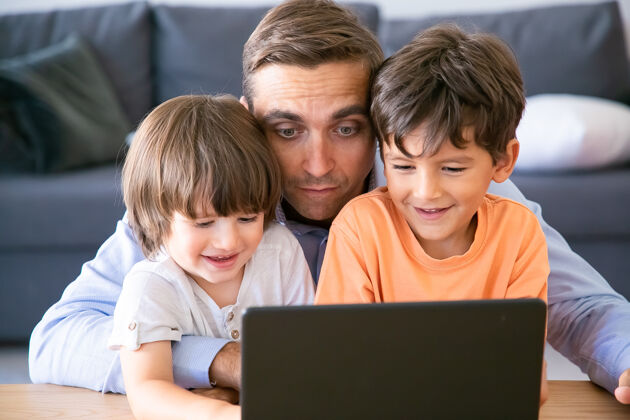 可爱中年爸爸抱着儿子 在笔记本电脑上看电影快乐的小男孩和爸爸坐在桌子旁 看着屏幕 微笑着父爱 童年和数字技术概念枕头室内爸爸