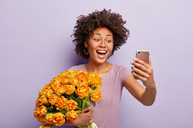 郁金香快乐的黑皮肤女士表达真挚的感情和情感 在社交网络上自拍分享照片 捧着漂亮的大束橙花 穿着休闲t恤 手机快乐高兴