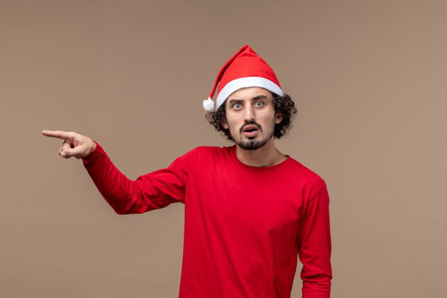 正面正面图棕色背景上穿着红色衣服 表情紧张的男性圣诞节帽子紧张微笑