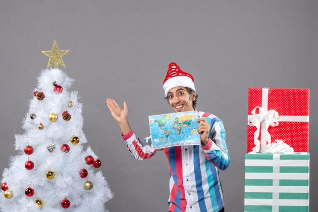 假期正面图微笑的男子戴着螺旋弹簧圣诞帽拿着世界地图指着圣诞树圣诞树印刷男人