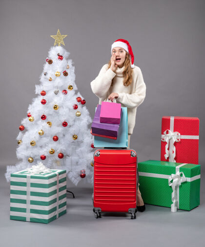 灰色正面图惊讶的金发女孩戴着圣诞帽 手里拿着红色的手提包和购物袋圣诞帽圣诞树盒子