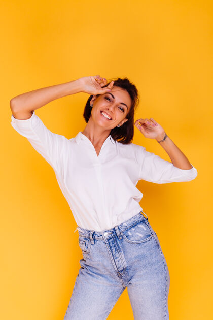姿势摄影棚拍摄的快乐女人短发穿着白衬衫和牛仔裤摆在黄色的墙上肖像情感脸
