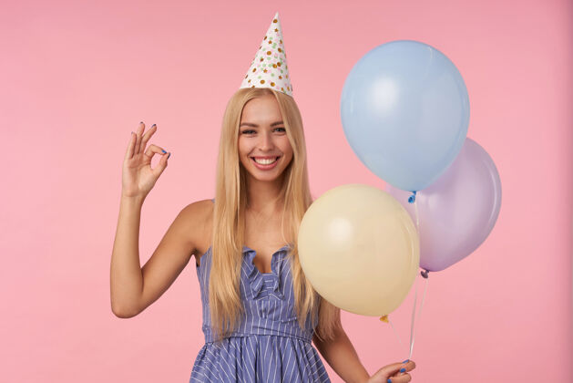 长发一幅欢快迷人的金发女性举手示意的肖像 站在粉色背景下 五颜六色的气球 开心地看着镜头 面带微笑女性五彩气球