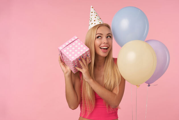 微笑快乐的年轻长发金发女士手持礼品包装盒 想知道里面是什么 和朋友们一起欢庆美好的聚会 站在粉色背景和五颜六色的气球上金发女士休闲