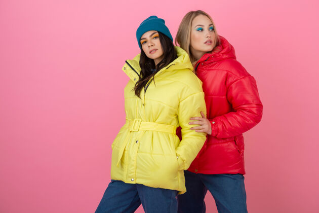 衣服两位迷人的时尚女性在粉色背景上合影 身着红黄相间的缤纷冬季羽绒服 暖衣时尚潮流朋友户外粉色