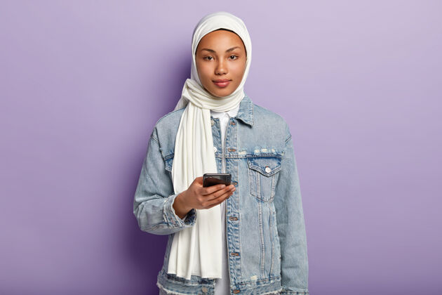 伊斯兰教一张美丽的阿拉伯女人的照片 皮肤黝黑健康 在现代手机上发短信 在帖子下面读评论 戴着白色头巾 穿着牛仔外套 模特们站在紫色的墙上宗教肖像阿拉伯语智能