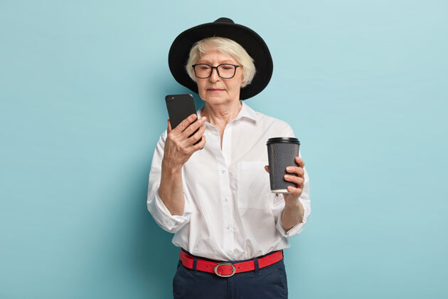 养老金严肃的老年妇女专注于智能手机设备 在互联网上搜索信息 在线阅读文章 喝咖啡去 退休人员使用简单的应用程序 穿着时尚的服装 在室内摆姿势风格短信腰带