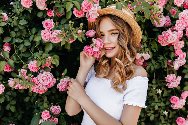 帽子快乐的白人女孩在大自然中摆姿势的户外写真一位悠闲的女士站在美丽的玫瑰丛旁 一头波浪形的头发树粉色花
