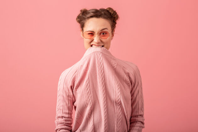 表情一个年轻漂亮的女人 穿着粉色毛衣 戴着墨镜 在粉色工作室的背景下扮鬼脸傻瓜漂亮鬼脸