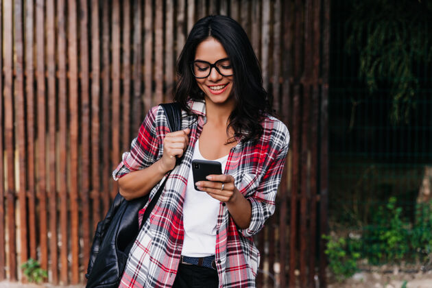 现代浪漫的棕褐色女人站在户外 背着背包和智能手机戴着眼镜的快乐拉丁女孩微笑着读着电话留言日光休闲女性