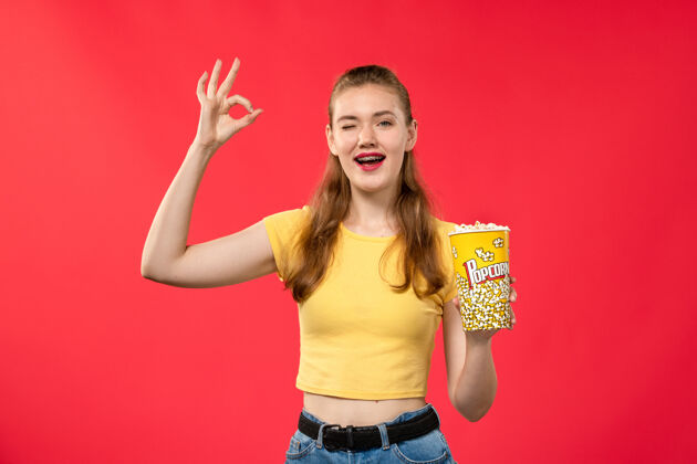年轻前视图年轻的女性在电影院拿着爆米花包 摆着姿势在浅红墙电影院的有趣的电影电影成人摆姿势