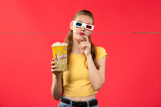 成人正面图年轻女性手持爆米花包 戴着d型太阳镜 在淡红色的墙上看电影年轻女性爆米花电影
