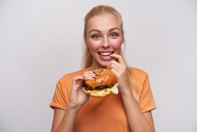 休闲快乐的蓝眼睛年轻可爱的金发女郎 随意的发型 兴奋地看着镜头 举起手拿着新鲜的汉堡包 食指放在腋下 笑容满面积极漂亮饮食
