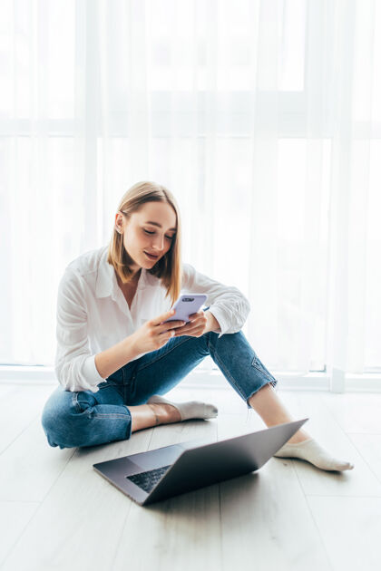 地板快乐的年轻女子坐在地板上 拿着笔记本电脑 微笑着在家里读手机短信或短信打字房子无线