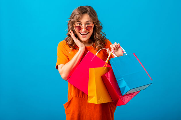 开朗图片中的兴奋迷人的微笑时尚女性购物狂穿着橙色的新潮连衣裙拿着购物袋在蓝色的工作室背景隔离情感持有微笑