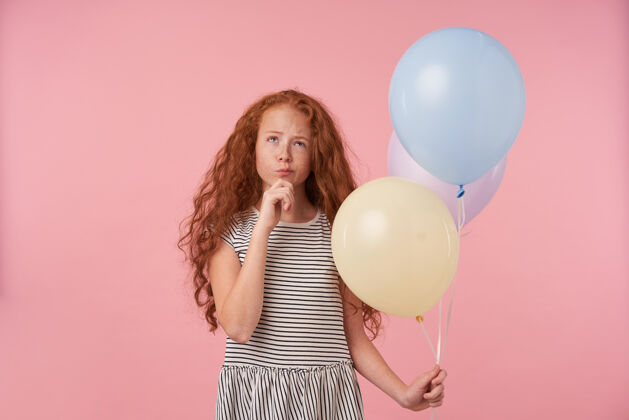 衣服沉思的红发卷发女孩儿的肖像 手里拿着气球 在粉色背景上摆姿势 手放在下巴上 若有所思地向上看 穿着休闲的条纹连衣裙雀斑手下巴