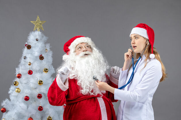 人们圣诞老人和女医生的前视图 女医生正在灰墙上检查他的健康状况圣诞老人服装圣诞老人