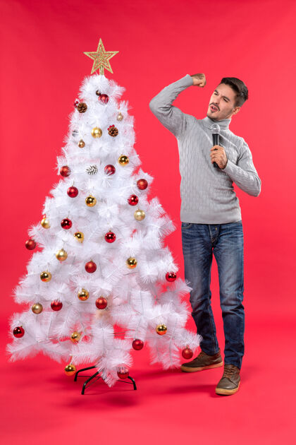 积极的家伙圣诞气氛积极的家伙穿着牛仔裤站在装饰圣诞树附近 拿着麦克风展示他的力量圣诞红色积极的