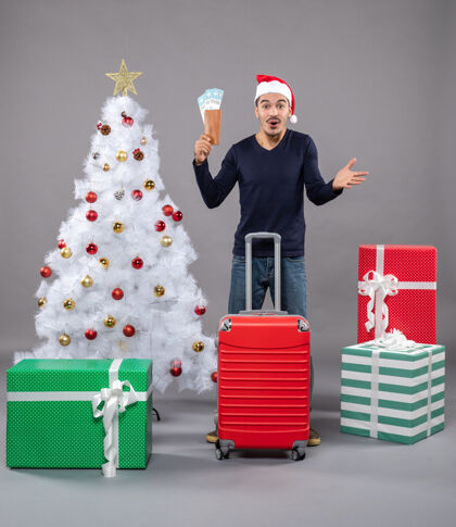 红色兴奋的年轻人拿着旅行票和红色手提箱围着圣诞树 礼物是灰色的购物圣诞圣诞树