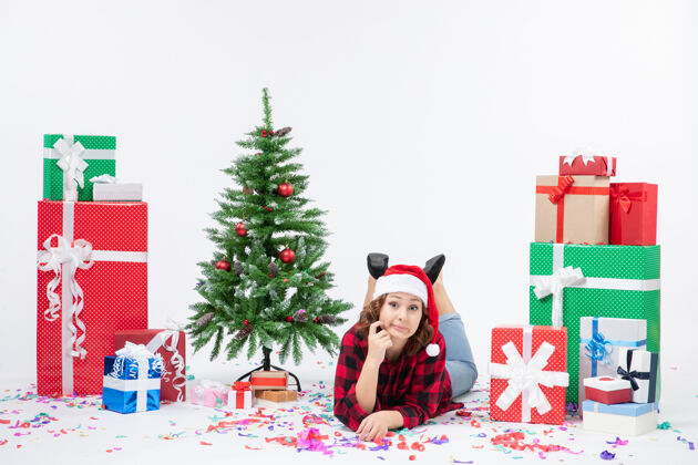 冬青正面图：年轻女子躺在圣诞礼物和小圣诞树周围 白色背景上 新年寒冷的女人 圣诞节下雪年轻女性礼物