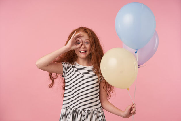 卷曲快乐可爱的红发卷发女孩儿举手 用ok手势对着眼睛 在粉色背景上用气球摆姿势 开心地看着镜头 笑容满面庆祝欧洲空气