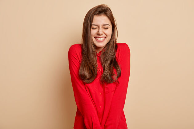 微笑积极的欧洲年轻女性的孤立镜头感觉享受和快乐 闭上眼睛 微笑广泛 穿着红色衣服 隔离在棕色的墙壁人 情感概念肖像深色头发满足