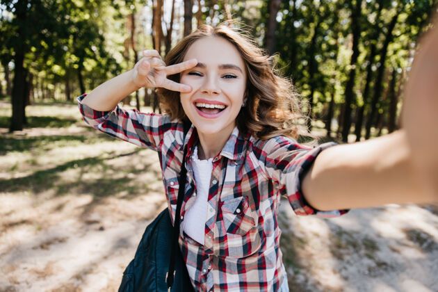 现代性感的白人女孩背着背包在森林里和和平标志合影美丽的卷发女人在公园里自拍和大笑的户外镜头游客金发公园