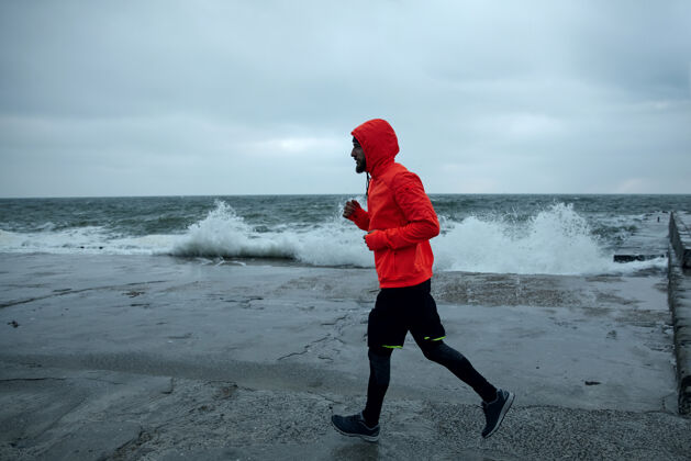 男户外拍摄的年轻运动员穿着温暖的运动服 在灰色和暴风雨的天气里在海边奔跑 注意力集中 双手保持在胸前水平黑发无遮蔽胡须