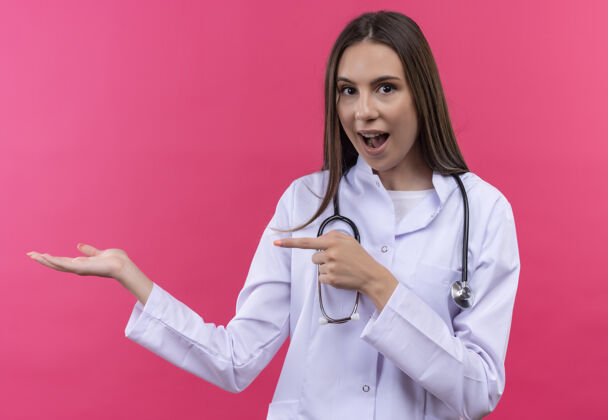 听诊器惊讶的年轻女医生穿着听诊器医用长袍指着手上孤立的粉红色背景点医生手