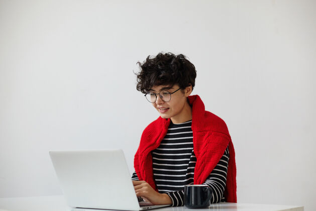 眼镜积极的年轻迷人的女人 短卷发 戴着眼镜 在现代办公室里用笔记本电脑工作 一边看屏幕一边打字 隔离在白色背景下笔记本电脑深色休闲