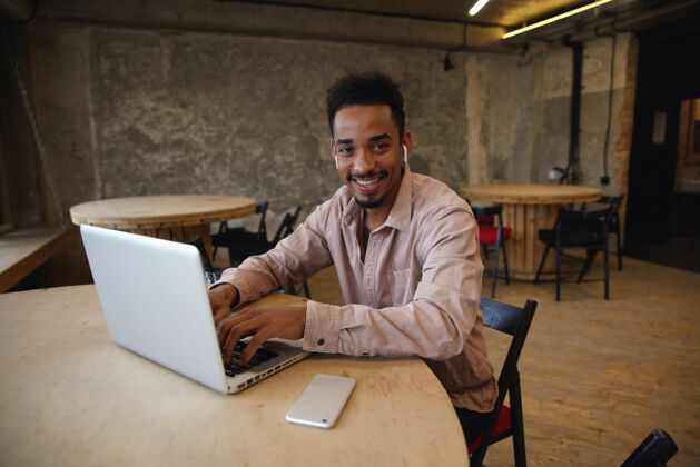 坐着快乐的年轻人 留着胡子 皮肤黝黑 穿着休闲服 坐在同事的座位上 面带迷人的笑容看着摄像机 手放在他现代笔记本电脑的键盘上非洲男性黑暗