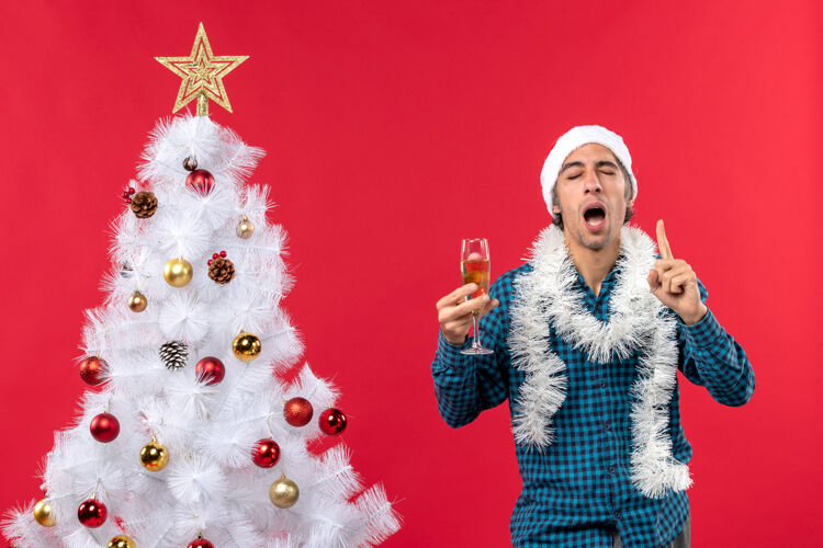 玻璃圣诞节心情自信坚定情绪化的年轻人戴着圣诞老人帽 穿着蓝色条纹衬衫 在圣诞树旁举起一杯葡萄酒人新年圣诞