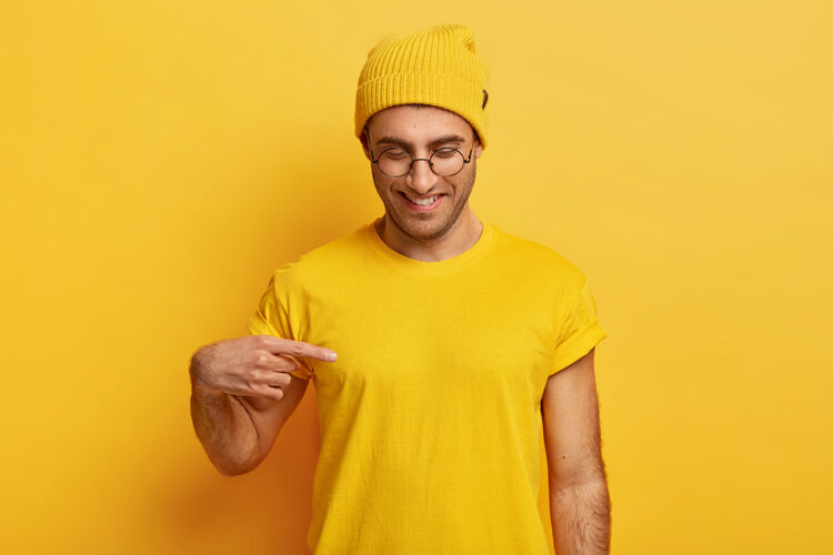 镜头积极的年轻男性指着t恤的空白处 展示你的设计或标志的空间 高兴地微笑 戴着眼镜 黄色的衣服 专注下来T恤高兴手指