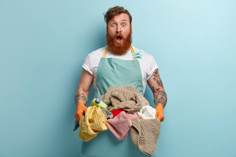 红发惊惶失措的红发大胡子男人穿着休闲的蓝色围裙 手里拿着一盆展开的衣服情绪化洗衣家伙