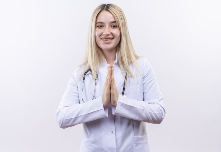 白人微笑的医生 年轻的金发女孩 戴着听诊器 穿着医用长袍 戴着牙套 在孤立的白色背景上做着祈祷的手势祈祷年轻穿着