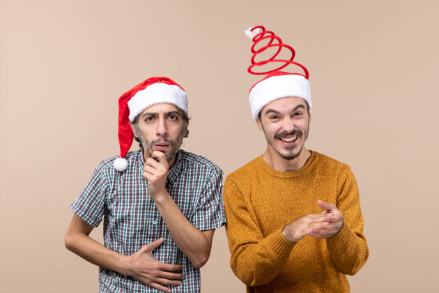 男人前视图两个家伙一个困惑和一个快乐的圣诞老人帽子米色孤立的背景米色男帽子