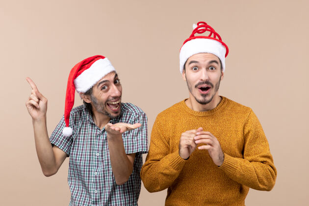 伙计们正面图两个戴着圣诞帽的家伙 一个在米色背景上展示另一个令人惊讶的东西头饰米色视图