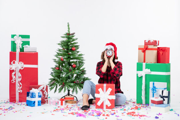 礼物前视图的年轻女子坐在圣诞礼物周围拿着白色墙上的时钟圣诞节节日时钟