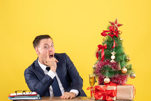 圣诞树坐在圣诞树旁桌子旁的一个好奇的人 把礼物放在黄色的桌子上好奇的男人办公室行政人员
