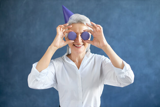 健康一个快乐的白发退休白人女性的横向肖像 她穿着白衬衫 戴着圆锥形帽子 眼睛里拿着两个圆形杏仁饼干聚会成人成熟