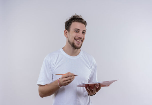 拿着微笑的年轻人穿着白色t恤 拿着铅笔和笔记本 背景是孤立的白色白色穿着笔记本