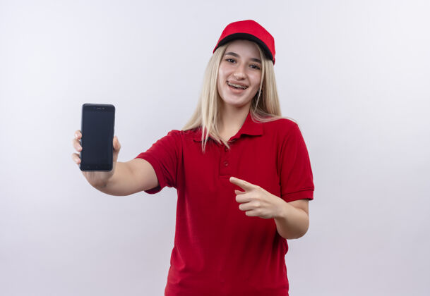 微笑微笑送货的年轻女孩穿着红色t恤 戴着帽子 戴着牙套 在孤立的白色背景下指着手里的手机T恤电话牙科