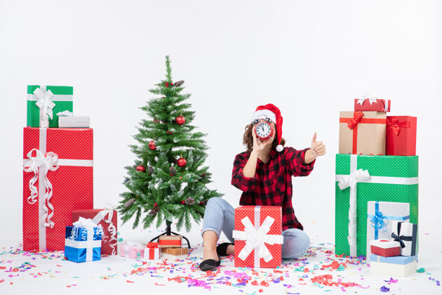 帽子前视图的年轻女子坐在圣诞礼物周围拿着白色墙上的时钟时钟圣诞节礼物