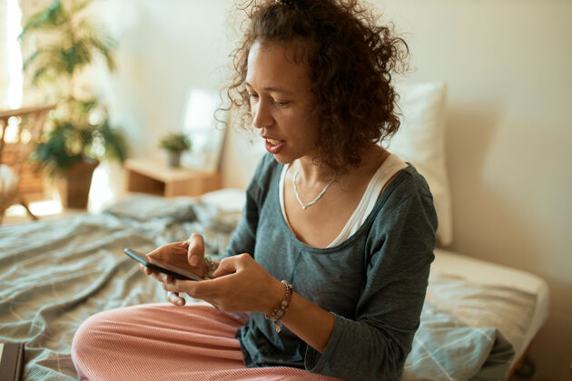 手机穿着随意的年轻拉丁女性自由职业者的画像 在网上卖商品 坐在床上用手机 打短信 和客户聊天坐着技术休闲
