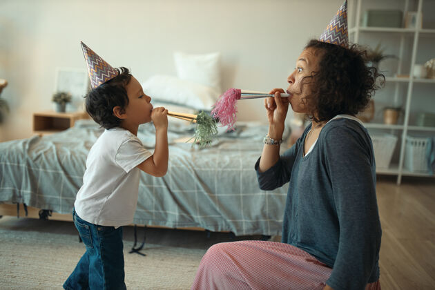 家在隔离检疫期间 一个人在家庆祝生日时 欢快的年轻混血女和她可爱的小儿子戴着锥形帽子吹口哨的侧视镜头帽子聚会哨子