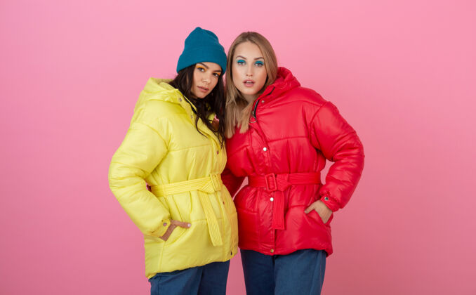 夹克两个迷人的女孩在粉红色的背景下 穿着鲜艳的红色和黄色的彩色冬季羽绒服摆姿势羽绒服女孩朋友